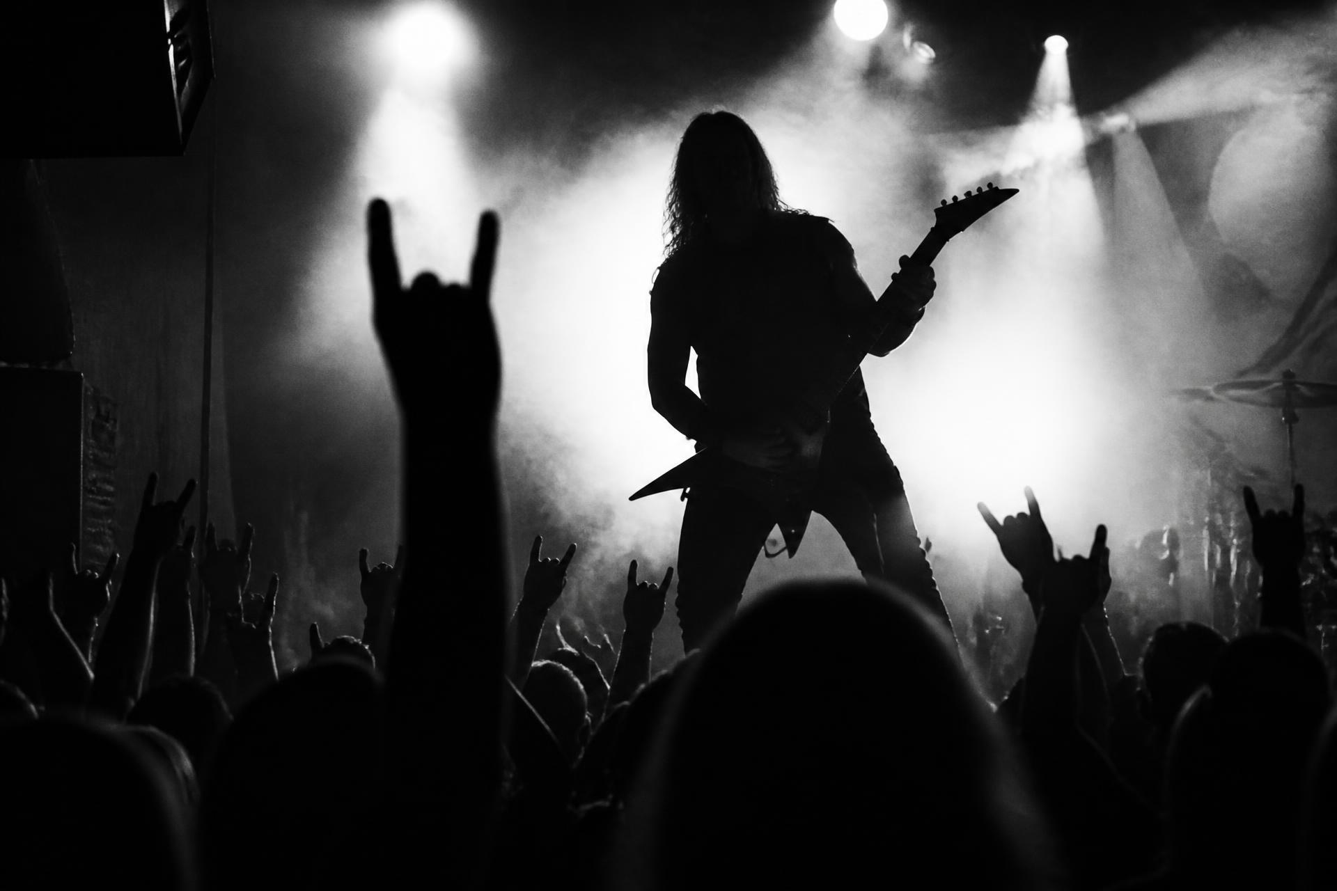 Metal guitarist performing to a raised-horns crowd under dramatic stage haze